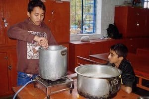 Two boys prepare food in a kitchen, with one stirring a large metal pot on a portable stove while the other watches beside a second cooking pot