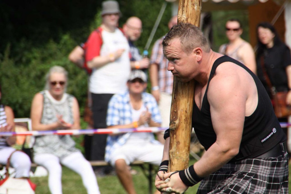 Strongman athlete in a kilt carrying a large wooden caber during a Scottish Highland games competition with spectators watching