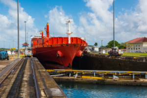 A bright red cargo ship navigating a lock in the Panama Canal, with rail tracks and tow locomotives guiding it. The lock gates are partially visible, with blue water below and buildings, trees, and a cloudy sky in the background.