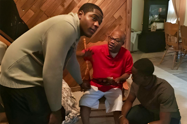 Nigeria GMS Leonard with kola nuts (600 x 400 px) Three men gather indoors, with an elderly man in a red shirt seated and holding a tray of kola nuts, engaging in conversation with two younger men beside him