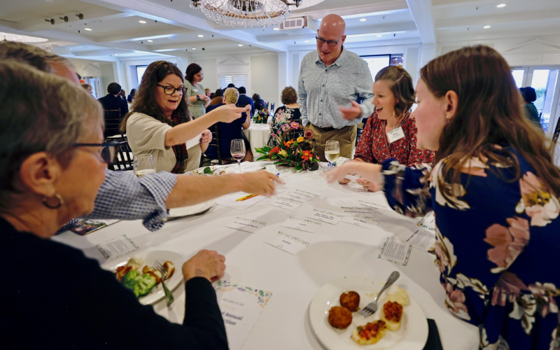 Guests seated around a round banquet table at a formal event enthusiastically sort small cards and chat over plates of hors d'oeuvres, wine glasses, and a floral centerpiece in a bright, chandelier-lit hall