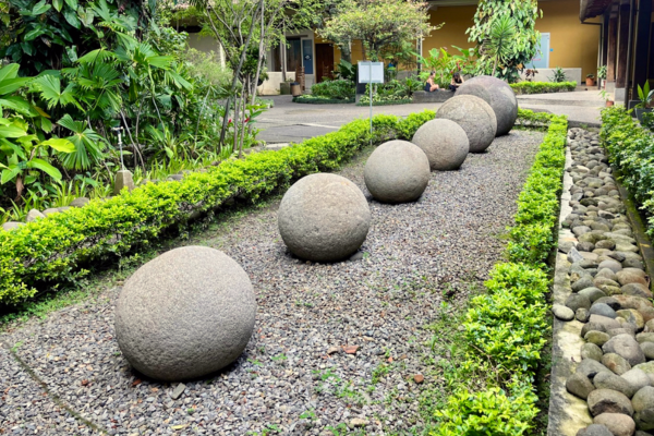 Row of pre-Columbian stone spheres of varying sizes displayed on a gravel path between manicured hedges in a verdant museum courtyard in Costa Rica