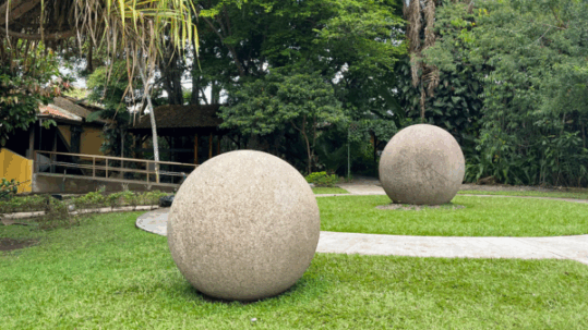 Two pre-Columbian stone spheres from Costa Rica rest on grass along a curving pathway in a lush tropical yard with trees and a rustic building