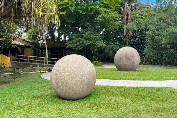 Two pre-Columbian stone spheres from Costa Rica rest on grass along a curving pathway in a lush tropical yard with trees and a rustic building