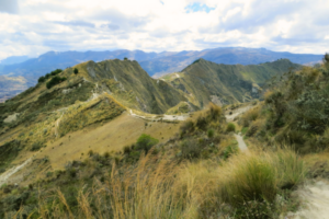 Mountain trail winding through the highlands of the Andes in Ecuador, surrounded by dry grass, rugged peaks, and dramatic cloud cover.
