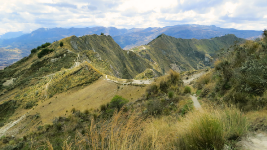 Mountain trail winding through the highlands of the Andes in Ecuador, surrounded by dry grass, rugged peaks, and dramatic cloud cover.