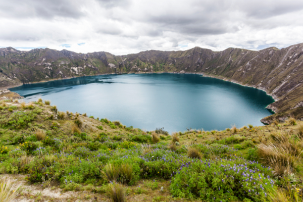 Crater lake surrounded by steep Andean mountains and lush highland vegetation under a cloudy sky, likely depicting Quilotoa in Ecuador.