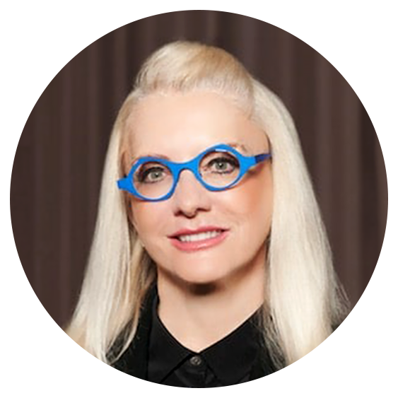 A woman with long blonde hair and bright blue glasses smiles at the camera against a neutral background