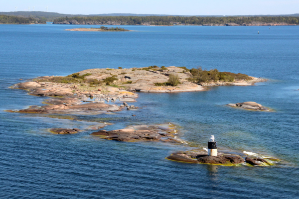 Rocky coastal islands with a small lighthouse surrounded by clear blue water and distant shoreline in the background