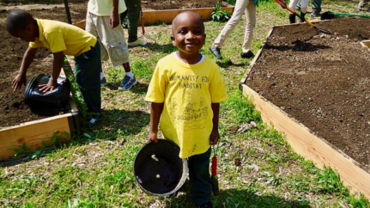 A smiling child holds a plant pot while others work in raised garden beds behind him