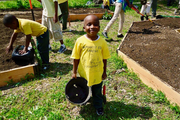 A smiling child holds a plant pot while others work in raised garden beds behind him