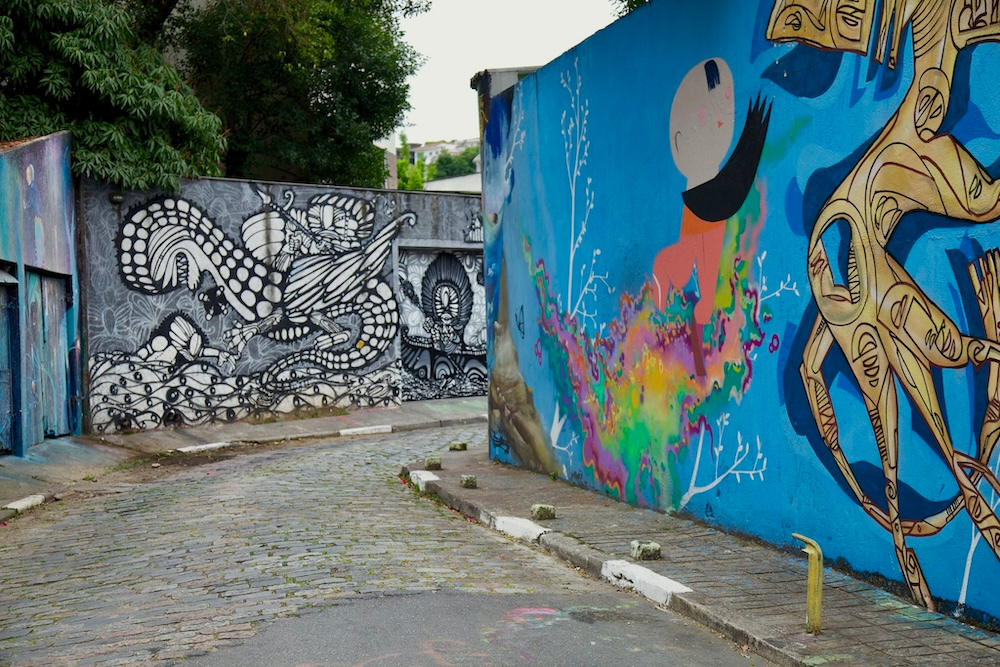 Cobblestone street bordered by colorful murals, including a blue wall with abstract figures and a black and white patterned wall under leafy trees