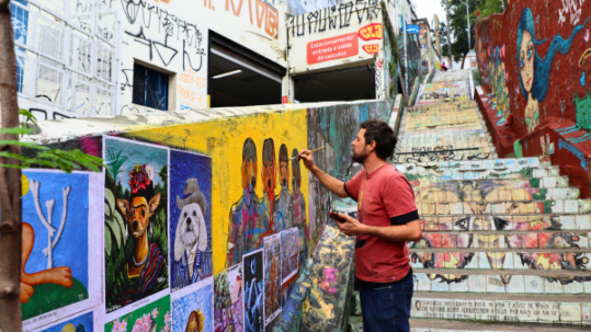 Man painting a wall mural beside graffiti-covered stairs and illustrated artwork panels in an urban street setting