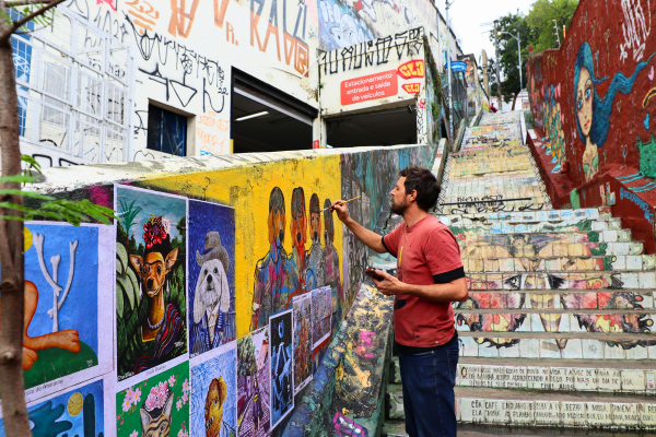 Man painting a wall mural beside graffiti-covered stairs and illustrated artwork panels in an urban street setting