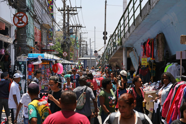 Crowded city market street with vendors, clothing stalls, overhead utility wires and pedestrians walking beneath an elevated walkway