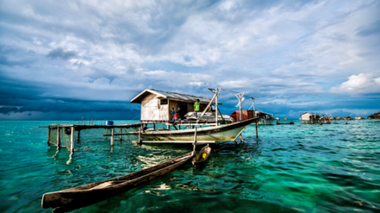 Wooden canoe beside a stilt house on bright turquoise sea under dramatic cloudy sky