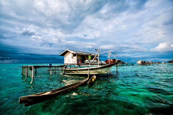 Wooden canoe beside a stilt house on bright turquoise sea under dramatic cloudy sky