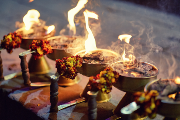 Lit brass oil lamps with flames, smoke and marigold flowers arranged on a ceremonial surface