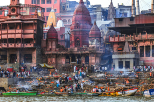 Riverside ghat with temples, boats, smoke and crowds gathered along the water in Varanasi, India