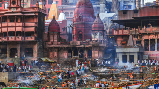 Riverside ghat with temples, boats, smoke and crowds gathered along the water in Varanasi, India
