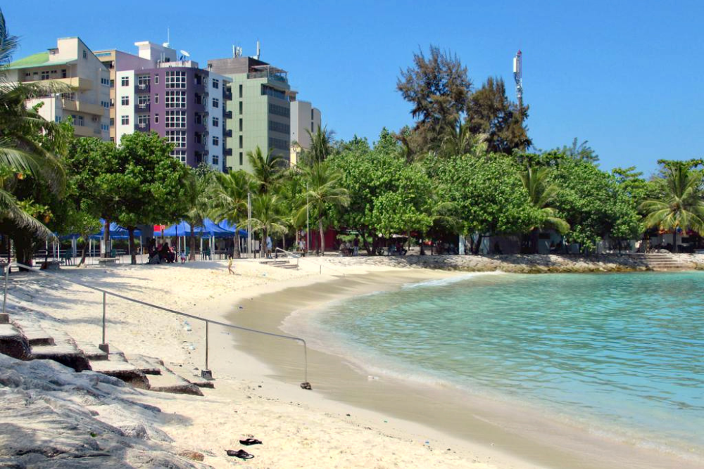 Tropical city beach with white sand, clear turquoise water, palm trees and colorful waterfront buildings under a bright blue sky