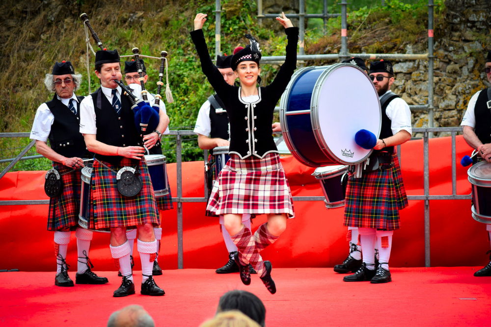 Scottish Highland dancer performing on a red stage with bagpipers and drummers in traditional tartan kilts at a cultural festival