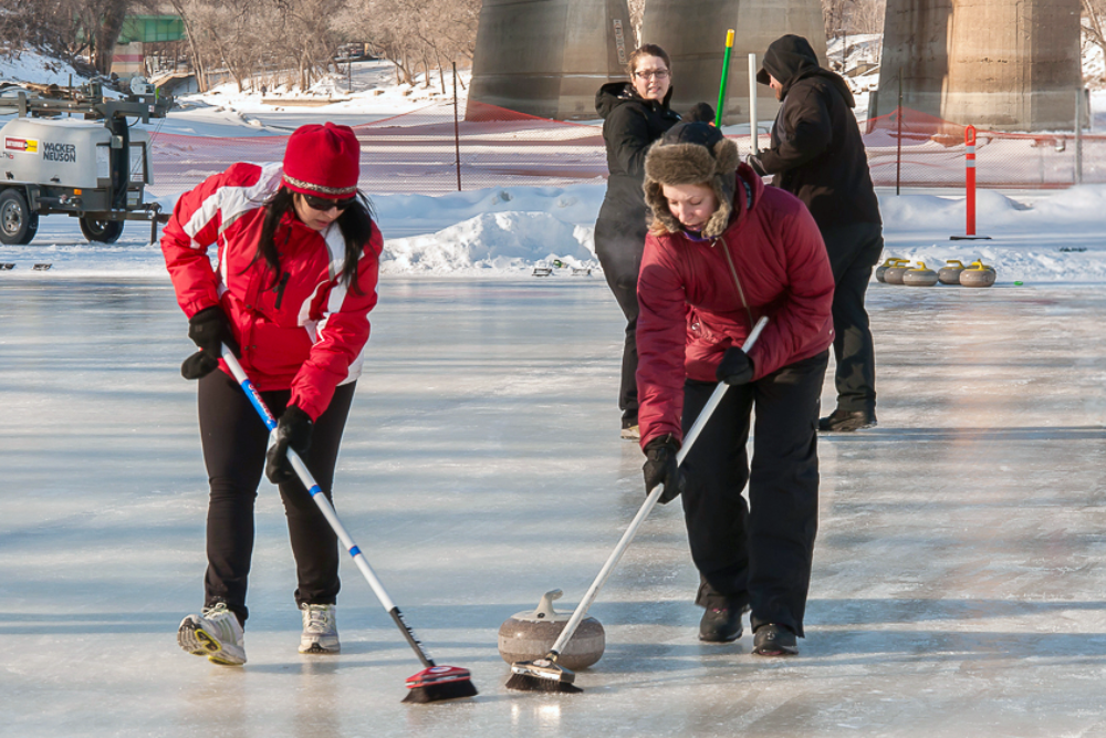Outdoor curling players using brooms to sweep a stone across a frozen ice rink surrounded by snow