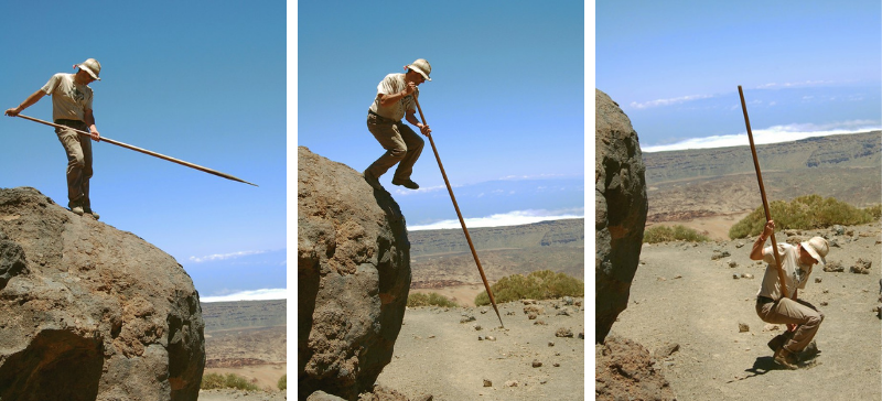 Man using a long wooden pole while balancing on and stepping down from a large desert boulder under a clear blue sky