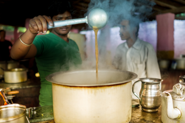 Man pouring hot tea from a ladle into a large steaming pot in a tea stall