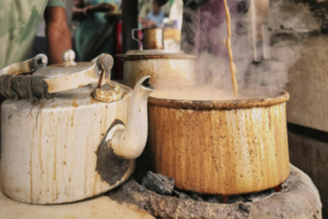 Steam rising from a stained metal teapot and large cooking pot on a stove as tea is poured into boiling liquid