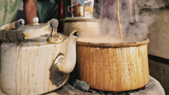 Steam rising from a stained metal teapot and large cooking pot on a stove as tea is poured into boiling liquid