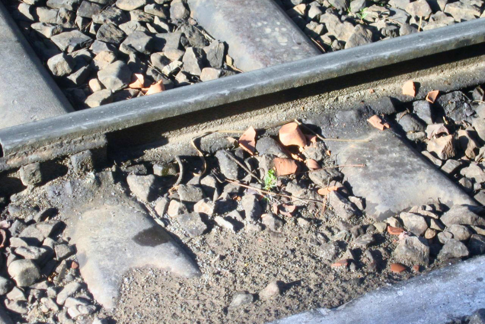 Railroad tracks with metal rails, wooden sleepers, gravel ballast and scattered broken tile pieces in bright sunlight