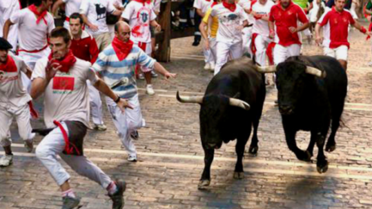 Two bulls charging along a cobblestone street as runners in white clothing with red scarves move alongside them during the Running of the Bulls festival