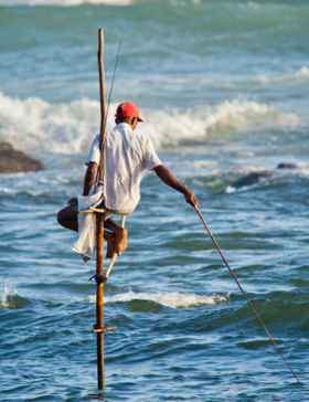 Stilt fisherman balancing on a wooden pole above ocean waves while holding a fishing rod
