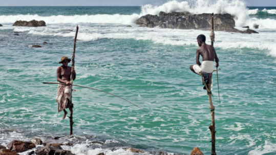 Two stilt fishermen sitting on wooden poles above rocky turquoise water with ocean waves behind them