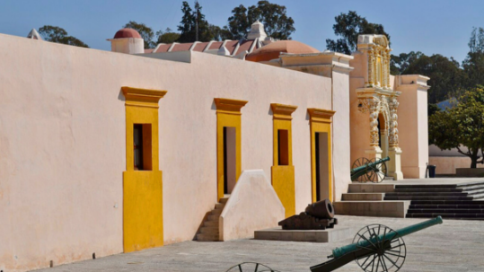 Courtyard of a historic fort with pastel-colored buildings, decorative architectural details, and several cannons displayed on wheeled carriages