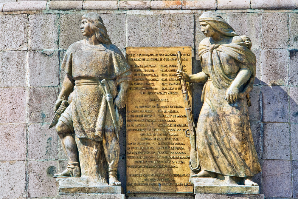 A stone monument featuring two sculpted figures in traditional clothing flanking a bronze commemorative plaque honoring participants in the Battle of Puebla, set against a large masonry wall