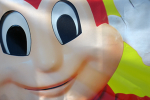 Close-up of the smiling face of the Jollibee mascot on a colorful sign, showing large cartoon eyes, rosy cheeks, and part of the red head against a bright background