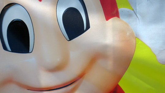 Close-up of the smiling face of the Jollibee mascot on a colorful sign, showing large cartoon eyes, rosy cheeks, and part of the red head against a bright background