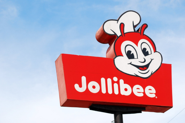 A roadside Jollibee restaurant sign featuring the smiling bee mascot wearing a chef's hat above a red rectangular sign with the white text "Jollibee." against a blue sky