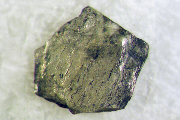Close-up of a rough, metallic gray crystal with reflective surfaces and irregular edges resting on a light-colored background