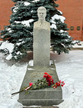 A stone memorial featuring a carved bust mounted on a tall granite pedestal, with several red flowers laid at its base. The monument stands in a snow-covered outdoor setting in front of an evergreen tree and a red brick building.