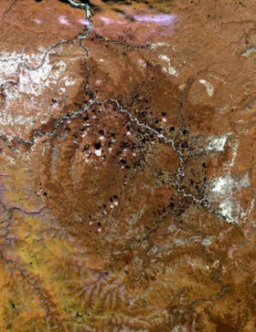 Satellite image of the Popigai impact crater in northern Siberia, showing a broad circular landform with branching river channels, rugged brown terrain, scattered dark lakes, and patches of white snow