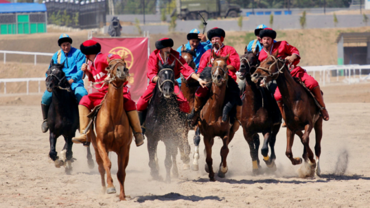 Kazakh horse riders in bright red and blue traditional clothing racing across a dusty field during an equestrian cultural event