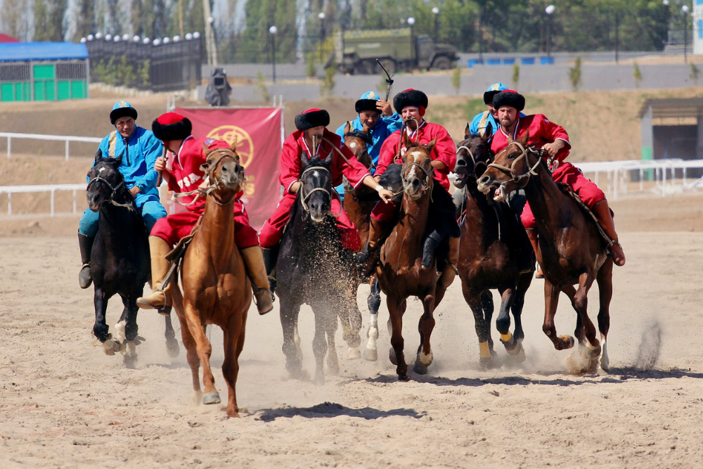 Kazakh horse riders in bright red and blue traditional clothing racing across a dusty field during an equestrian cultural event
