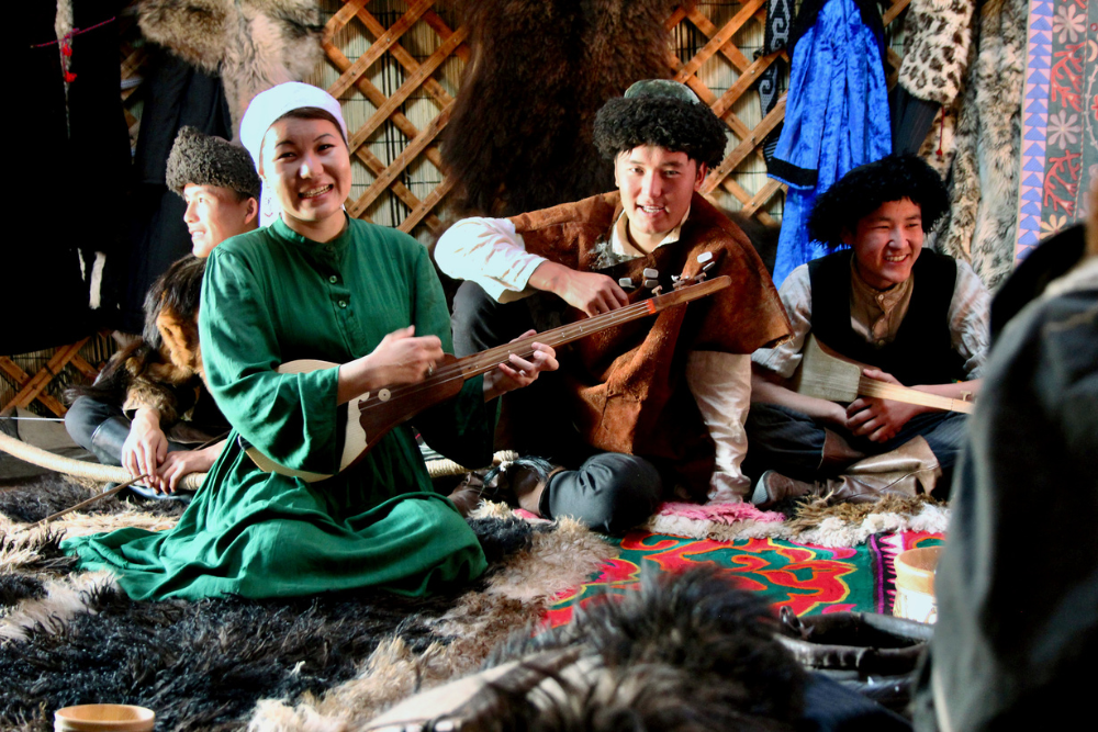 Kazakh musicians in traditional clothing playing string instruments inside a decorated yurt lined with furs and colorful textiles