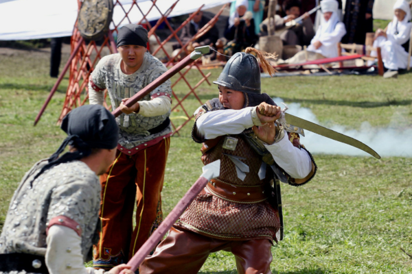 Kazakh warriors in traditional armor reenacting a sword fight on a grassy field beside a yurt during a cultural festival