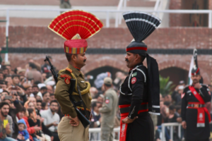 Two ceremonial border guards from India and Pakistan stand face-to-face in decorated military uniforms with distinctive fan-shaped headdresses during the Wagah border flag-lowering ceremony, surrounded by spectators