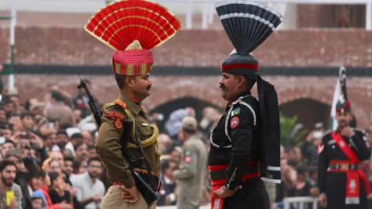 Two ceremonial border guards from India and Pakistan stand face-to-face in decorated military uniforms with distinctive fan-shaped headdresses during the Wagah border flag-lowering ceremony, surrounded by spectators