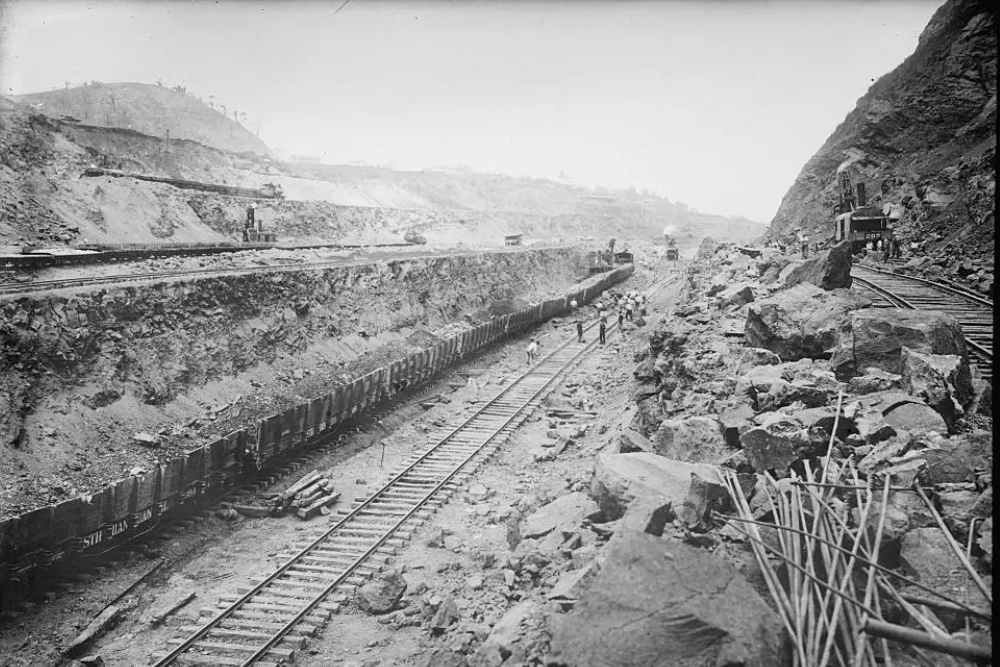 A historic black-and-white photograph showing railroad construction through a rocky mountain cut, with multiple railway tracks, freight cars, steam locomotives, workers, and excavation equipment spread across the rugged landscape